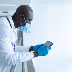 African american male doctor in hospital corridor wearing face mask using tablet. medical professional at work during coronavirus covid 19 pandemic.